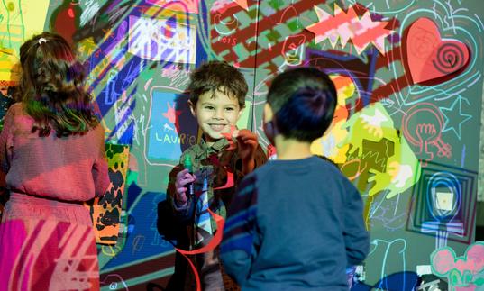 a young boy plays with his friend up against a wall projected with the work of Jean Michel Basquiat. They are both smiling and having a really nice time