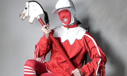 Gazelle Twin wearing a jester costume with a hobby horse, seated on a chair