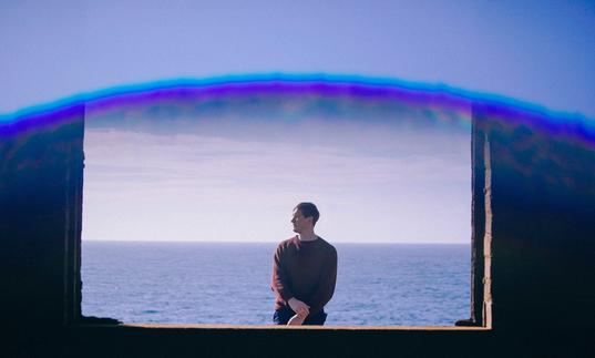 Erland Cooper standing in front of the sea, as seen through a window