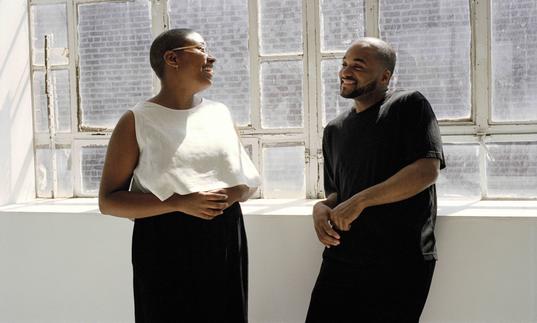 Cecile McLorin Salvant and Sullivan Fortner leaning on a ledge in front of windows with sun streaming in