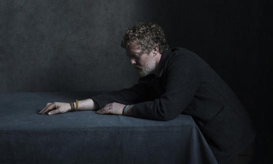 Glen Hansard stretching his right arm across a table. He is wearing leather bracelets.