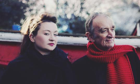 Eliza and Martin Carthy sitting in a boat outdoors. Eliza is looking at the camera and Martin is wearing a red scarf.