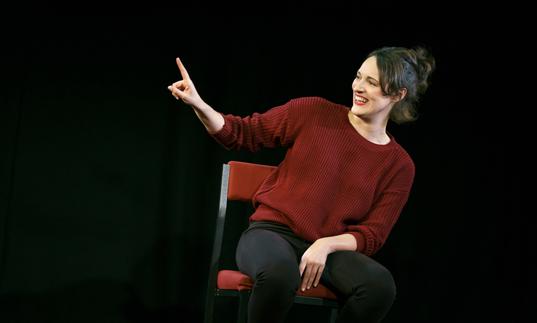Phoebe Waller-Bridge sitting on a chair in a dark space with her arm and finger pointing up
