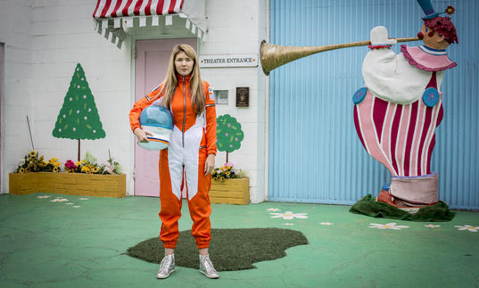 Beatie Wolfe wearing an orange space suit, with a colourful house behind her