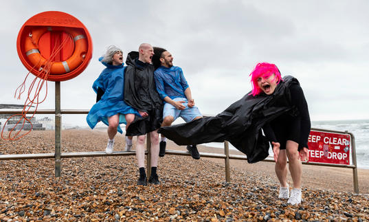 4 people in anoraks are on a windy beach. One has a bright pink wig.