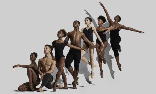 seven dancers are in a line in serveal balletic poses. The positions range from low to the ground, to the dancer at the back of the line in a jump.