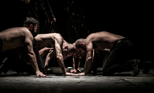 four topless men in a dark room appear to be eating from the floor