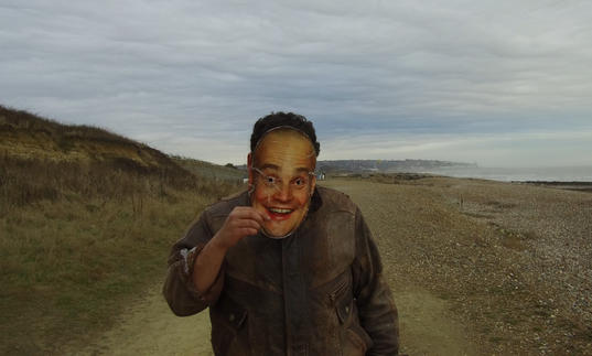 A man iin a green field holds a mask of a face over his own face