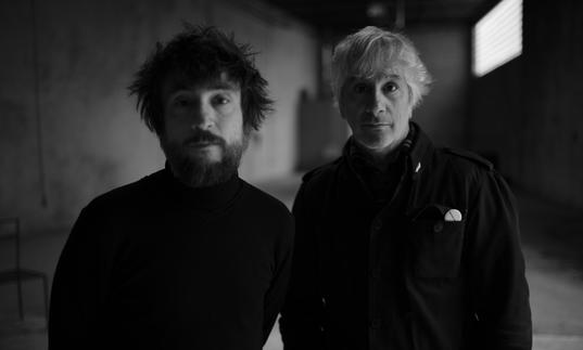 A black and white photo of Lee Ranaldo and Raul Refree in an empty concrete room, wearing black shirts