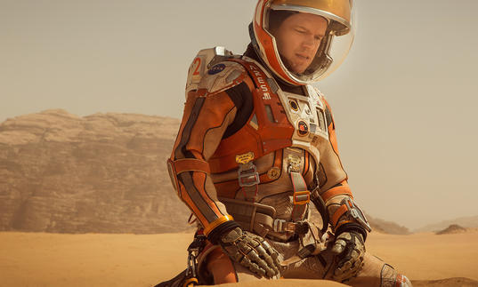Matt Damon in an orange space suit on a dusty, desert-style landscape in The Martian