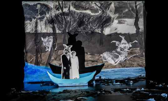 A figure that is half-man, half-kangaroo is standing in a boat with Fleur Elise Nobel's unnamed character. They look like figures in a pop-up book, against a paper painted backdrop and vivid blue colour in the water. Their shadows are prevalent against the paper backdrop.  