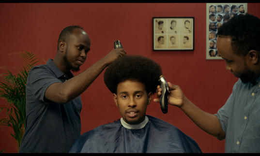 a man sitting in the barber chair with two barbers cutting his hair