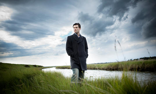 An image of Ryan standing on the grassy river bank, a dramatic grey sky overhead 