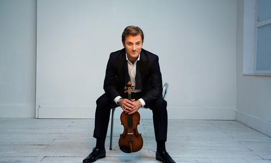 Renaud sitting on a chair in a white room with a cheeky grin on his face, clutching his violin