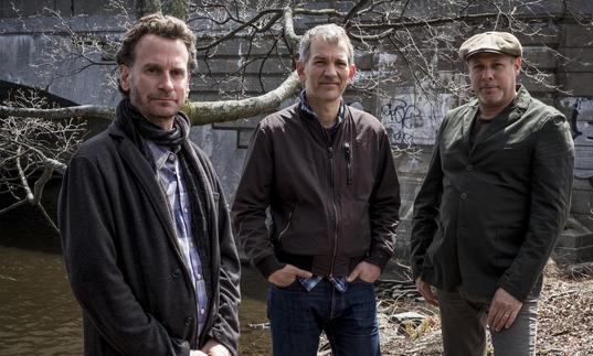 the trio stand next to a canal. They are dressed casually and it looks like autumn/winter