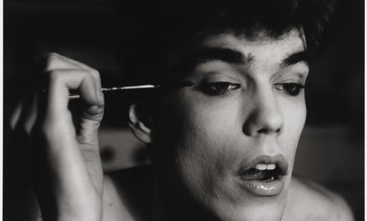 An black and white image of a man open mouthed, as he concentrates on putting on mascara