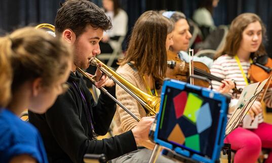 A group of young people in a orchestra, one boy is playing the trumpet and two girls in the background are playing the violin.
