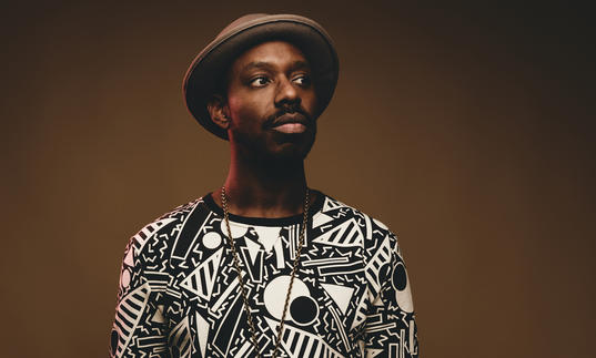 Shabaka Hutchings wearing a patterned top and hat, looking to his left.