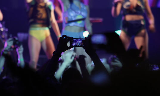 a person in a crowd holds their phone up to film the girl band on stage