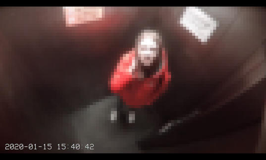 A pixelated view from a CCTV camera of someone looking up at the camera in a small room. They are wearing a red coat and they have brown hair.