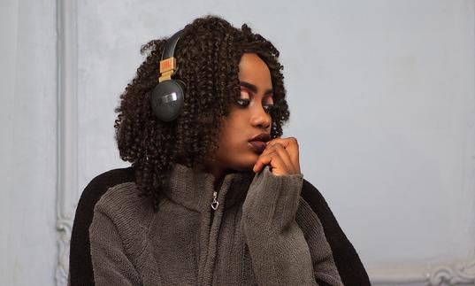 A young black woman with brown curly hair sits on the floor with headphones on looking into the distance. She's wearing a grey jumper, black tights and white trainers.