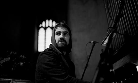 black and white image of Kit Downes playing the organ with an arched church window in the background