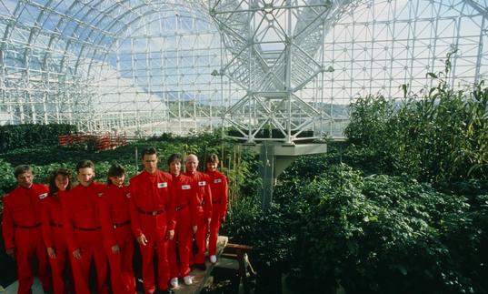A large group of people dressed in red jumpsuits stand inside a large glass biodome, with plants