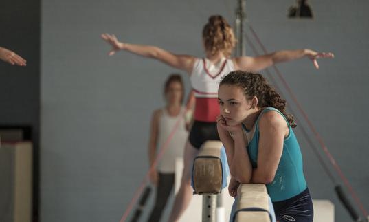 teenage girl looks wistful leaning on a balancing bar in a gymnastics hall