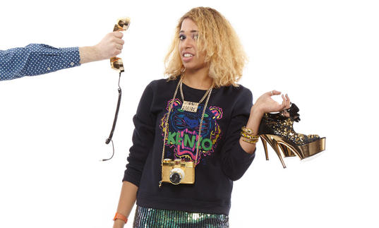 Paula Varjack holding stiletto shoes, looking at the camera unimpressed as someone holds up a phone with cut-off cord to her