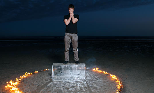 David Finnegan stands on an ice block, in a desert. A ring of fire burns around him.
