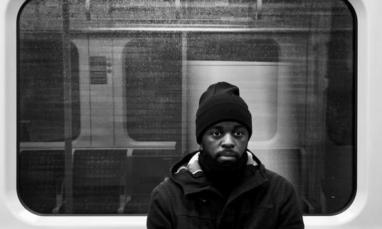 black and white photo of Alpha Mist sitting in a tube carriage, he is wearing a big coat and a beanie hat, with a neutral expression