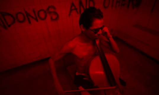 a man plays a cello in a dark room full of red light and graffiti on the wall