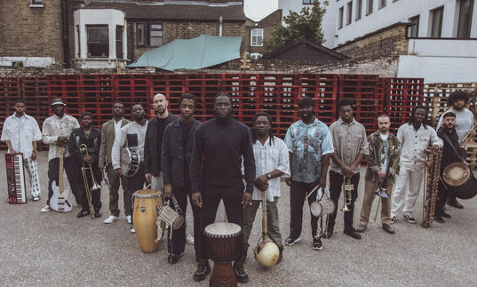 Balimaya Project standing in a triange formation with their instruments at their feet, in front of stacked red crates.
