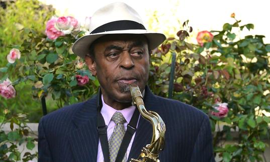 Archie Shepp playing a saxophone. He is wearing a black suit with a white hat and standing in front of a rose bush.