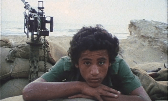 A young man lies on sand bags with a automatic gun behind him