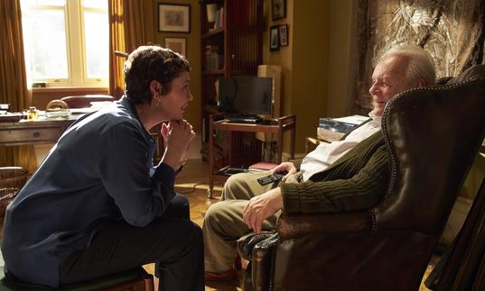 a daughter and her father sit looking at each other in a cluttered living room