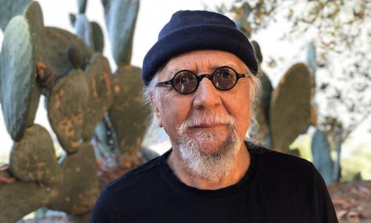 Charles Lloyd in a black beanie hat and small round sunglasses, standing in front of a large cactus