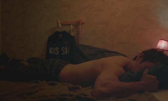 A man lies in on a bed belly down, in a dimly lit room, with a Russia hoodie in the background