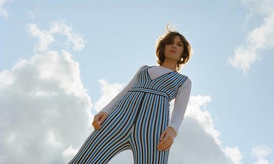 colour photo of Aldous Harding against a blue sky background. Photo is shot from below and she wears a blue striped jumpsuit