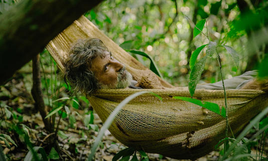 A man with grey hair and a beard lies in a hammock in foliage, in a still from El Father Plays Himself