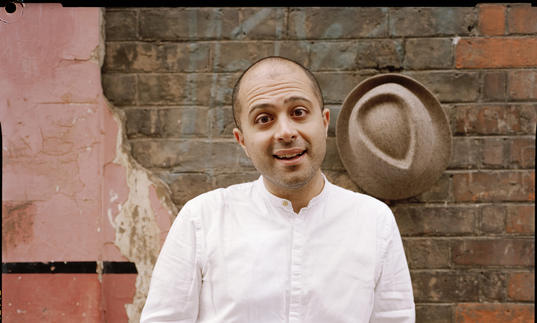 Mahan Esfahani smiling in front of a wall, with his hat hanging on the wall behind him