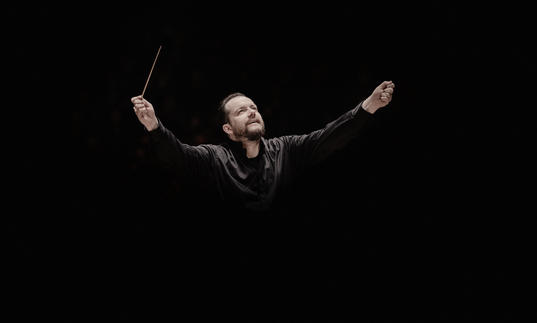 Andris Nelsons with his arms raised. He's holding a conductor's baton in his right arm, and he appears to be coming out of the black background. 