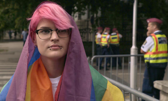 Tobi, a young person with pink hair, is draped in the rainbow flag in front of a few police officers in a still from Colors of Tobi