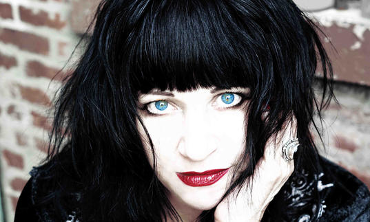 A portrait image of musician Lydia Lunch, with black hair and blue eyes