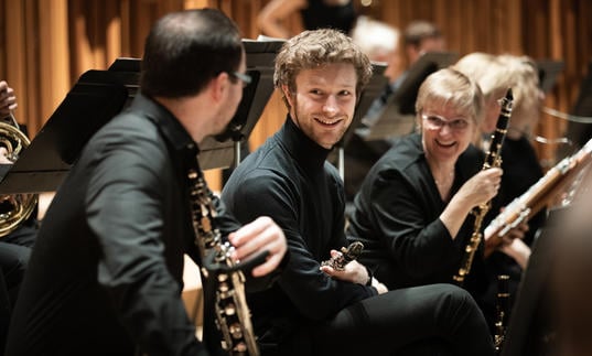 Two members of Britten Sinfonia smile at another member, who has his back to the camera. They are all on the Barbican stage, holding their instruments.