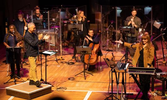 Hannah Peel performing with Paraorchestra. From behind a keyboard on the left of the stage she is gesturing to the ensemble who are applauding.