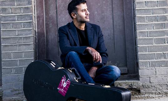 Hamza Namira sitting in a doorway with his guitar case on the floor in front of him.