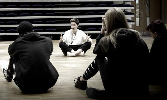 A black and white image of a theatre workshop with an instructor facing participants