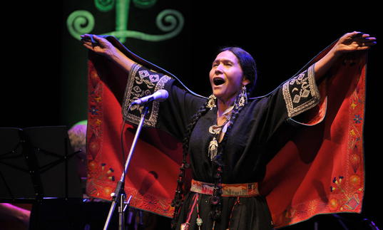 Luzmila Carpio singing, she is wearing a colourful traditional indigenous outfit with her hands outstretched above her