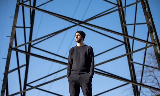 a colour photo of Manu Delago standing in front of an electricity pylon and a blue sky